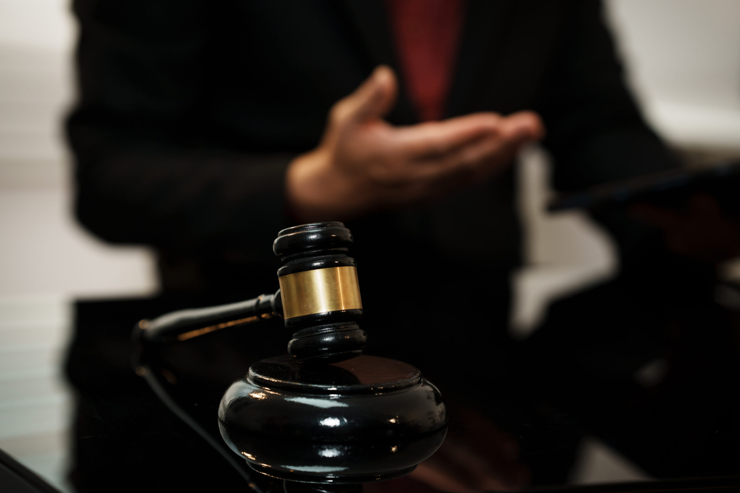 Close-up of judge gavel on desk with blurred lawyer gesturing in background, courtroom or legal office setting, shallow depth of field.